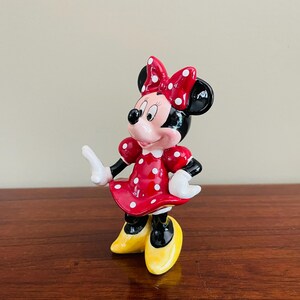 May include: A Minnie Mouse figurine. The figure is wearing a red dress with white polka dots, a matching bow, and yellow shoes. The figure has black ears and a pink face. The figurine is standing on a wooden surface.