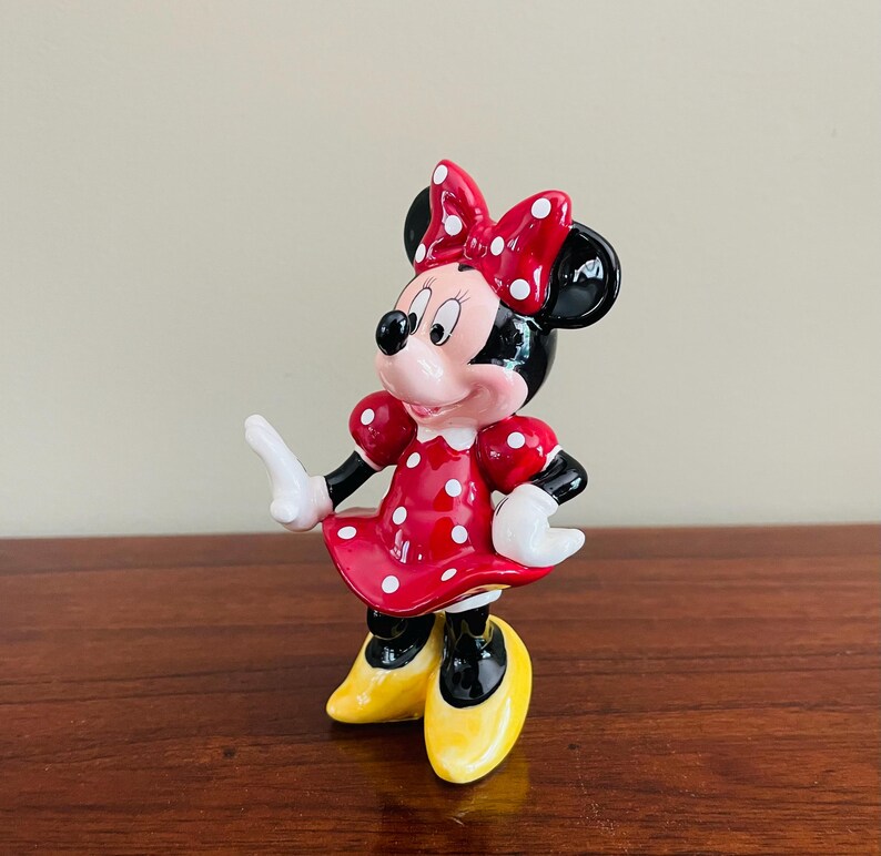 May include: A Minnie Mouse figurine. The figure is wearing a red dress with white polka dots, a matching bow, and yellow shoes. The figure has black ears and a pink face. The figurine is standing on a wooden surface.