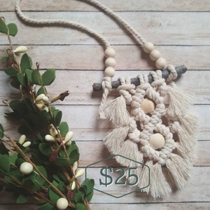 Może przedstawiać: Naszyjnik z makramą z drewnianymi koralikami i małą gałązką. Naszyjnik jest biały i ma frędzle na dole. Cena wynosi 25 €.