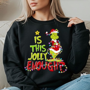 May include: A black long-sleeve sweatshirt featuring a graphic of a green character wearing a red Santa hat and suit, wrapped in colourful Christmas lights. The text reads IS THIS JOLLY ENOUGH? in green and red letters with a yellow star above.