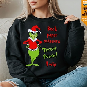 May include: A black long-sleeve sweatshirt featuring a graphic of a green character wearing a red Santa hat and red shirt. The text reads: Rock paper scissors, Throat Punch! I win. The character has a smirk and hands on hips.
