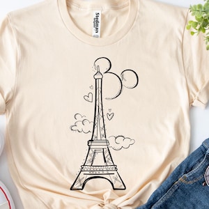 K&ouml;nnte beinhalten: Ein cremefarbenes T-Shirt mit einer schwarzen Strichzeichnung des Eiffelturms, Ballons, Wolken und Herzen. Das Shirt wird mit wei&szlig;en Turnschuhen, Jeansshorts, Sonnenbrille und einem Lutscher kombiniert.