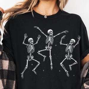 May include: A black t-shirt featuring a white graphic print of three dancing skeletons surrounded by small stars. The shirt is worn with an unbuttoned black and beige plaid flannel shirt over it, creating a layered look with the dark fabric.