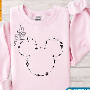 May include: A pale pink sweatshirt with a black outline of Mickey Mouse's head made of stars and a fairy. The sweatshirt has long sleeves and a crew neck. The text "Personalisation Available" is on the bottom right.