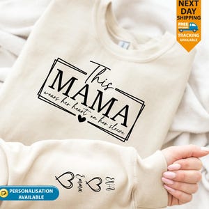 May include: A cream-coloured sweatshirt with the text "This MAMA wears her heart on her sleeve" in black. The sleeve features heart graphics and names. The sweatshirt has a crew neck and is made of a soft material.