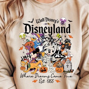 May include: Beige sweatshirt with a Halloween-themed Disneyland graphic. The design features Mickey Mouse as a mummy, Donald Duck, Goofy, Pluto, and other Disney characters in Halloween costumes. Text reads "Walt Disney's Disneyland" and "Where Dreams Come True Est. 1955."