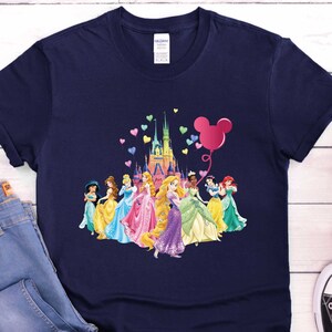 May include: Navy blue t-shirt featuring a colorful graphic of Disney princesses, a castle, and a Mickey Mouse balloon. The princesses are in their iconic gowns. The shirt is paired with blue jeans and black and white sneakers.