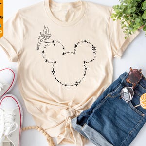 May include: A cream-coloured t-shirt with a black outline of Tinkerbell and a Mickey Mouse head made of stars. The shirt is styled with blue denim shorts, white trainers, and accessories. The image includes text: "NEXT DAY SHIPPING" and "PERSONALISATION AVAILABLE."