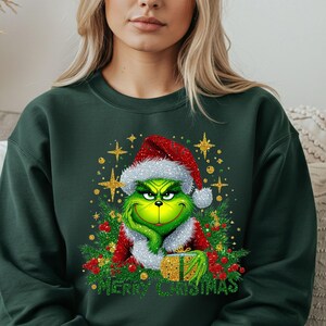 May include: Dark green sweatshirt with a Grinch graphic wearing a Santa hat and holding a gift. The design includes the text "Merry Christmas" in white. The sweatshirt is a festive Christmas item.