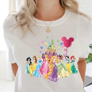 May include: White t-shirt featuring a colorful graphic of Disney princesses in front of a castle, with a pink Mickey Mouse balloon and heart-shaped balloons. The princesses are wearing gowns in various colors.