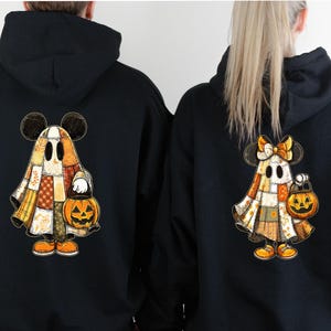 May include: Black hooded sweatshirts with a Halloween design. The design shows a cartoon ghost character with Mickey Mouse ears, holding a jack-o'-lantern. The ghost is wearing a patchwork quilt-style outfit. The character is wearing orange shoes.