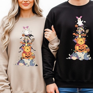 May include: Beige and black sweatshirts with a Halloween-themed graphic. The graphic shows Winnie the Pooh characters in costumes, including a ghost, witch, and pumpkin. The sweatshirts are long-sleeved.