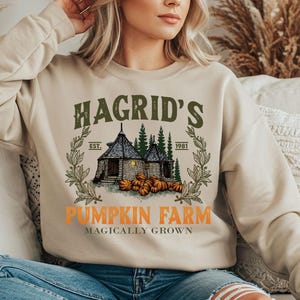 May include: Beige sweatshirt featuring the text "Hagrid's Pumpkin Farm" in green and orange. The design includes an illustration of a small house, pumpkins, and trees. The sweatshirt is a neutral colour.