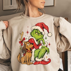 May include: A beige crewneck sweatshirt featuring a graphic print of a green character wearing a red Santa hat and suit, sitting next to a brown dog with reindeer antlers and a red bow. The design includes red and green star accents.
