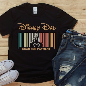 Pode incluir: Camiseta preta com um gr&aacute;fico de estilo retro que diz "Disney Dad" com um c&oacute;digo de barras e um castelo de desenho animado. O c&oacute;digo de barras diz "Scan for Payment".
