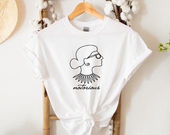 May include: White t-shirt with a black line drawing of a woman wearing glasses and a necklace. The text "notorious" is printed below the drawing.