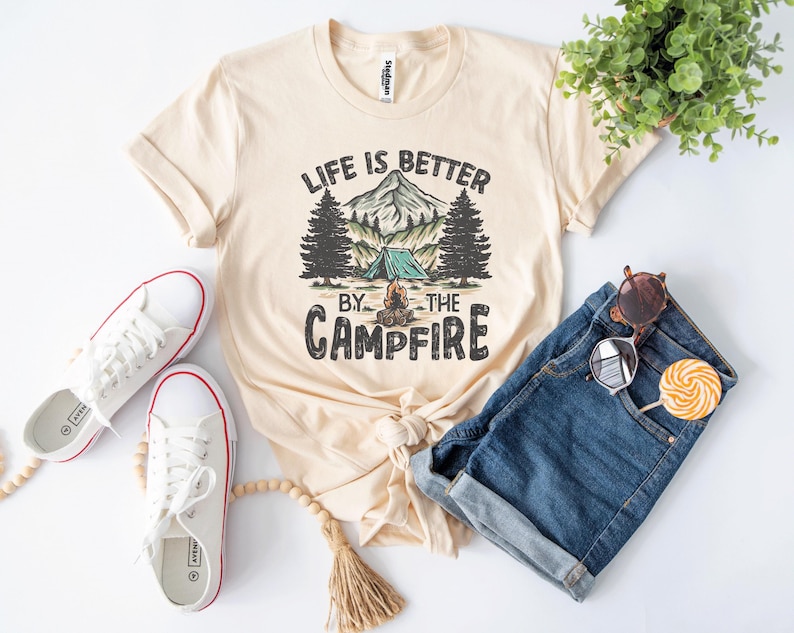 May include: Cream-colored t-shirt with the text "Life is Better by the Campfire" and a graphic of a mountain, trees, and a tent. The shirt is paired with denim shorts, white sneakers, and sunglasses.
