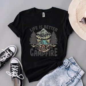 May include: Black t-shirt with a camping graphic featuring a mountain, trees, and a campfire, with the text "LIFE IS BETTER BY THE CAMPFIRE." The shirt is paired with black sneakers, blue jeans, and a straw hat.
