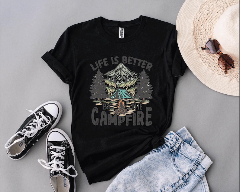 May include: Black t-shirt with a camping graphic featuring a mountain, trees, and a campfire, with the text "LIFE IS BETTER BY THE CAMPFIRE." The shirt is paired with black sneakers, blue jeans, and a straw hat.