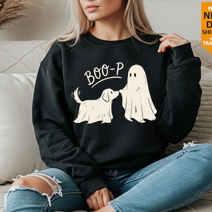 May include: A black long-sleeve sweatshirt featuring a graphic print of a dog and a ghost standing together. Above the figures, the text reads BOO-P in a white, curved font. The design is centred on the chest of the garment.