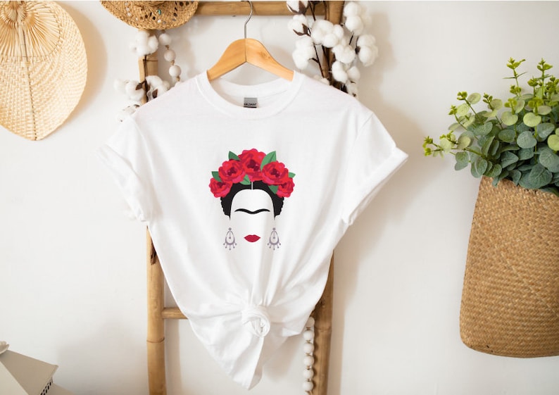 May include: White t-shirt with a black and white illustration of Frida Kahlo wearing a red flower crown.