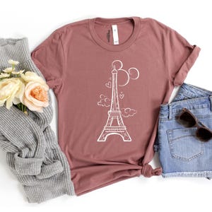 K&ouml;nnte beinhalten: Ein mauvefarbenes T-Shirt mit einem wei&szlig;en Eiffelturm-Motiv, Herzen und Wolken. Das Shirt wird mit Jeansshorts und Sonnenbrille kombiniert. Ein grauer Schal und Blumen sind zur Seite angeordnet.