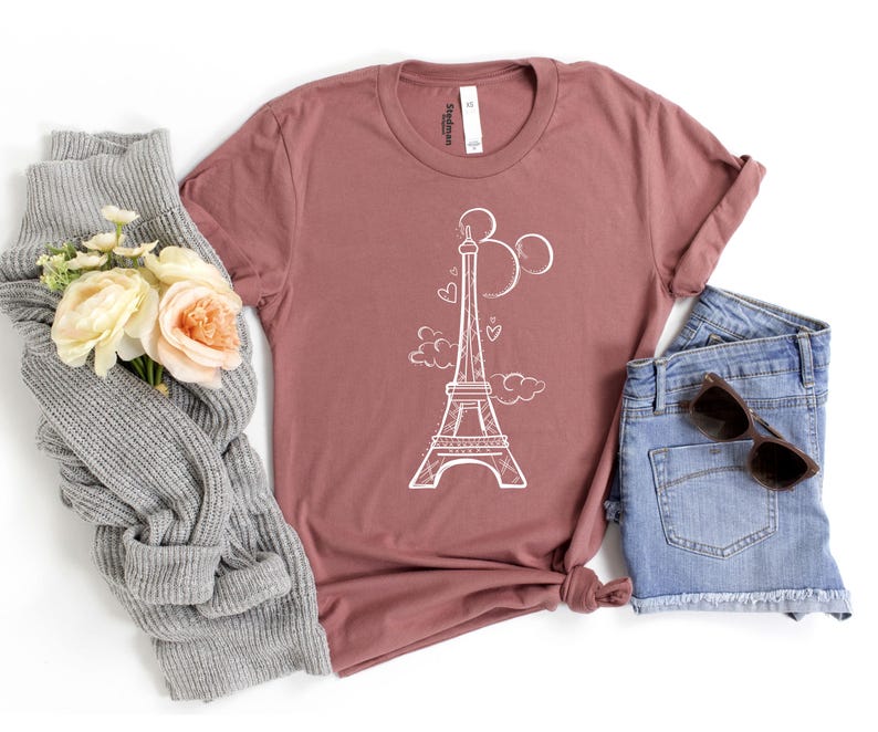 K&ouml;nnte beinhalten: Ein mauvefarbenes T-Shirt mit einem wei&szlig;en Eiffelturm-Motiv, Herzen und Wolken. Das Shirt wird mit Jeansshorts und Sonnenbrille kombiniert. Ein grauer Schal und Blumen sind zur Seite angeordnet.