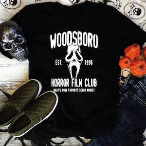 May include: Black t-shirt with a white graphic design featuring a ghost face mask and the text "Woodsboro Est. 1996 Horror Film Club What's your favourite scary movie?"