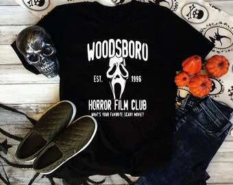 Pode incluir: Camiseta preta com um design gráfico branco que apresenta uma máscara de fantasma e o texto "Woodsboro Est. 1996 Horror Film Club What's your favorite scary movie?"