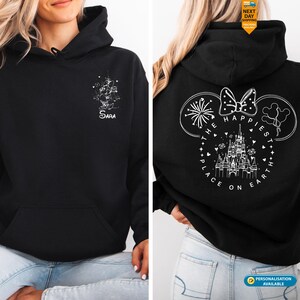 May include: Black hooded sweatshirt with a front pocket. The front features a white graphic and the name "Sara". The back displays a white design of Mickey Mouse ears, a bow, fireworks, and a castle with the text "The Happiest Place on Earth".