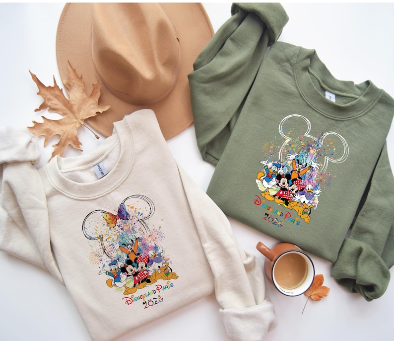 May include: Two sweatshirts, one cream and one olive green, with a colourful Disney character design. The design includes Mickey Mouse ears and the text "Disneyland Paris 2023". A tan hat and autumn leaves are also in the image.