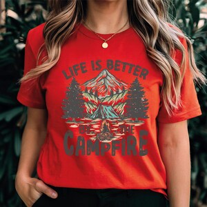 May include: Red t-shirt with a camping graphic. The design features a mountain, trees, a tent, and a campfire, with the text "LIFE IS BETTER BY THE CAMPFIRE" in white and green.
