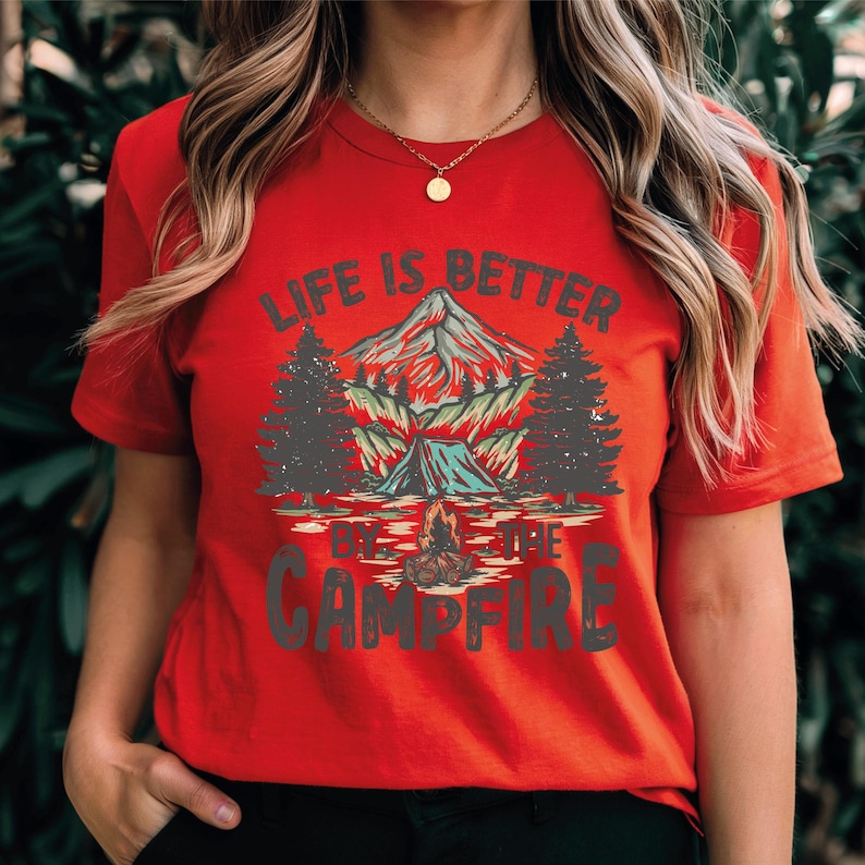 May include: Red t-shirt with a camping graphic. The design features a mountain, trees, a tent, and a campfire, with the text "LIFE IS BETTER BY THE CAMPFIRE" in white and green.