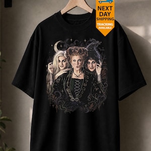 May include: A black short-sleeved t-shirt featuring a graphic print of three women with distinct hairstyles and dark clothing. The central figure has red hair, while the others have blonde and black hair. The design includes a crescent moon and stars.