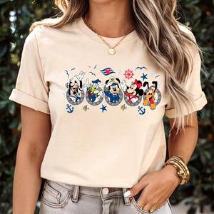 Pu&ograve; includere: T-shirt beige con un motivo nautico con personaggi dei cartoni animati in obl&ograve;. Il design include Topolino e Minnie, Paperino, Pippo e Pluto, insieme ad ancore, stelle e un timone.