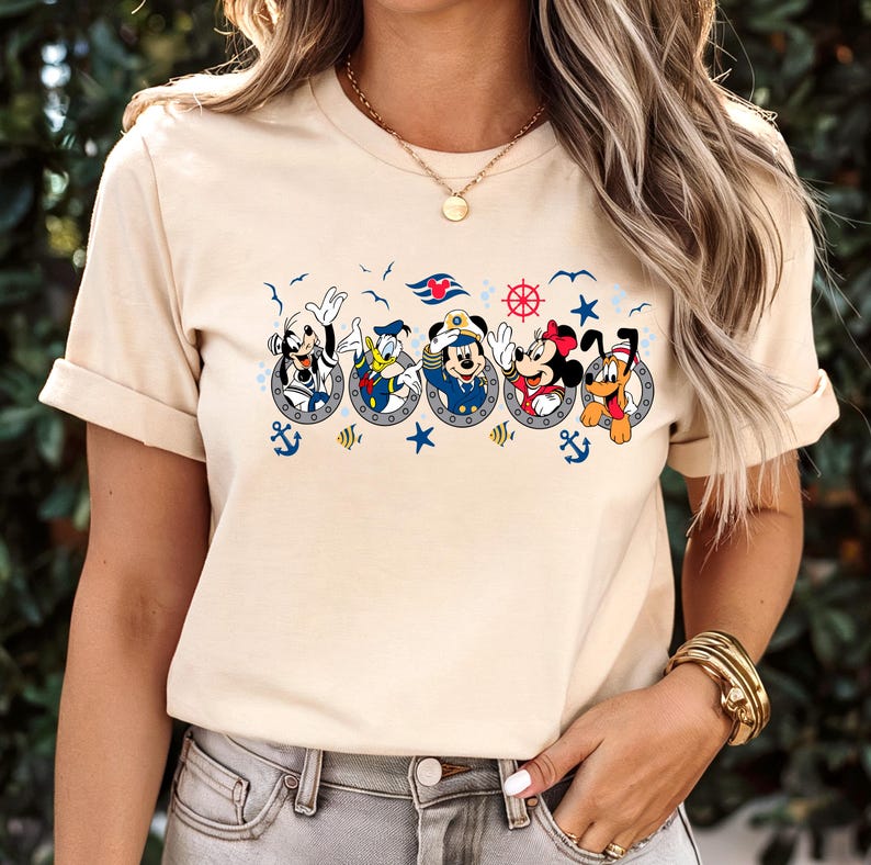 Pu&ograve; includere: T-shirt beige con un motivo nautico con personaggi dei cartoni animati in obl&ograve;. Il design include Topolino e Minnie, Paperino, Pippo e Pluto, insieme ad ancore, stelle e un timone.