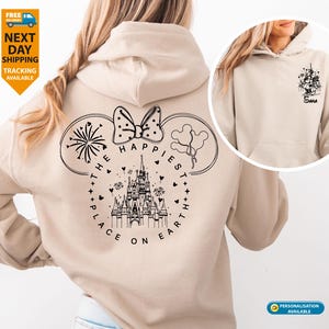 May include: Beige hoodie with a black graphic on the back featuring a castle, bow, fireworks, and balloons, with the text "THE HAPPIEST PLACE ON EARTH." A smaller graphic is on the front. The hoodie has a drawstring hood and a front pocket.