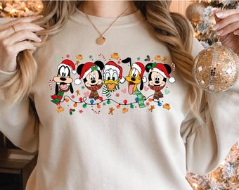 May include: Beige sweatshirt featuring a festive design with cartoon characters. The graphic includes Mickey Mouse, Minnie Mouse, Donald Duck, Goofy, and Pluto, all wearing Santa hats and scarves, with Christmas lights and gingerbread men.