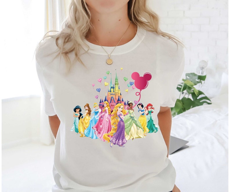 May include: White t-shirt featuring a colorful graphic of Disney princesses in front of a castle, with a pink Mickey Mouse balloon and heart-shaped balloons. The princesses are wearing gowns in various colors.