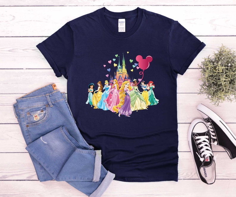 May include: Navy blue t-shirt featuring a colorful graphic of Disney princesses, a castle, and a Mickey Mouse balloon. The princesses are in their iconic gowns. The shirt is paired with blue jeans and black and white sneakers.