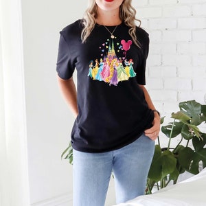 May include: Black t-shirt featuring a colorful graphic of a castle, princesses, and a Mickey Mouse balloon. The design includes a variety of colors, including pink, yellow, blue, and purple. The shirt is paired with blue jeans.