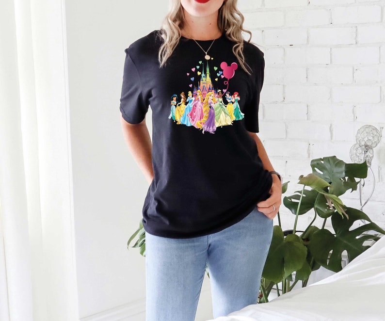 May include: Black t-shirt featuring a colorful graphic of a castle, princesses, and a Mickey Mouse balloon. The design includes a variety of colors, including pink, yellow, blue, and purple. The shirt is paired with blue jeans.