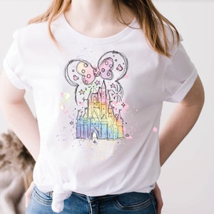 May include: A white t-shirt with a colorful illustration of Cinderella's castle and a Mickey Mouse bow. The illustration is surrounded by a watercolor-like effect with pink, blue, and yellow splatters.