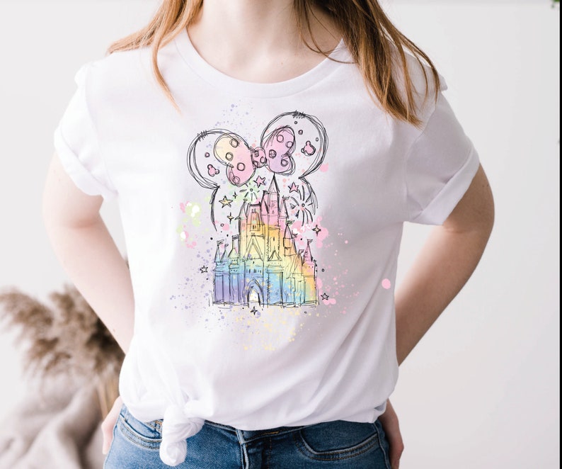 May include: A white t-shirt with a colorful illustration of Cinderella's castle and a Mickey Mouse bow. The illustration is surrounded by a watercolor-like effect with pink, blue, and yellow splatters.