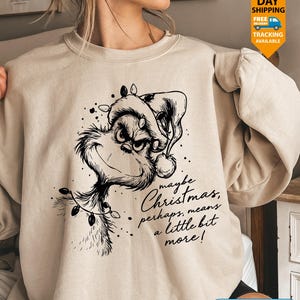 May include: A beige crewneck sweatshirt featuring a black line art illustration of a grumpy character wearing a Santa hat and wrapped in string lights. The text reads, maybe Christmas, perhaps, means a little bit more! in a cursive script.