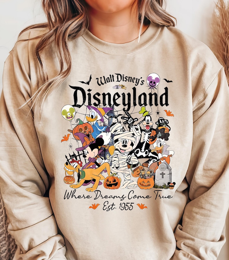 May include: Beige sweatshirt with a Halloween-themed Disneyland graphic. The design features Mickey Mouse as a mummy, Donald Duck, Goofy, Pluto, and other Disney characters in Halloween costumes. Text reads "Walt Disney's Disneyland" and "Where Dreams Come True Est. 1955."