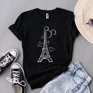 K&ouml;nnte beinhalten: Schwarzes T-Shirt mit wei&szlig;er Grafik des Eiffelturms, Herzen und Wolken. Dazu blaue Jeansshorts, schwarze Sneaker, ein Strohhut und eine Sonnenbrille. Ein l&auml;ssiges Outfit zum Thema Reisen.