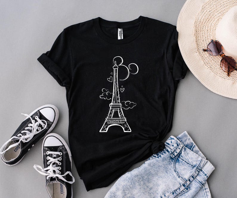 K&ouml;nnte beinhalten: Schwarzes T-Shirt mit wei&szlig;er Grafik des Eiffelturms, Herzen und Wolken. Dazu blaue Jeansshorts, schwarze Sneaker, ein Strohhut und eine Sonnenbrille. Ein l&auml;ssiges Outfit zum Thema Reisen.