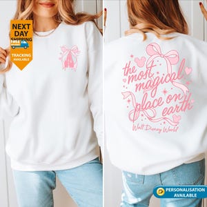 May include: White sweatshirt with a pink castle and bow design on the front. The back features the text "the most magical place on earth" with a bow and hearts. Walt Disney World is printed below the text.
