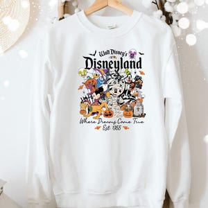 May include: White crew neck sweatshirt with a Halloween-themed Disney graphic. The design features various Disney characters dressed in costumes, including Mickey Mouse and Donald Duck. The text reads "Walt Disney's Disneyland" and "Where Dreams Come True Est. 1955".